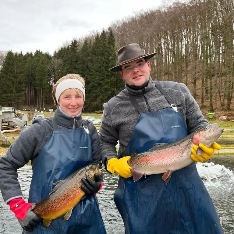 Bettina und Christian Grasse stehen in Wathosen am Teich und halten zwei große Regenbogenforellen.